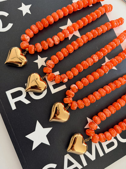 Four beaded necklaces with orange coral-like round beads and shiny gold heart charms, arranged diagonally on a black card with white stars and partial white lettering.