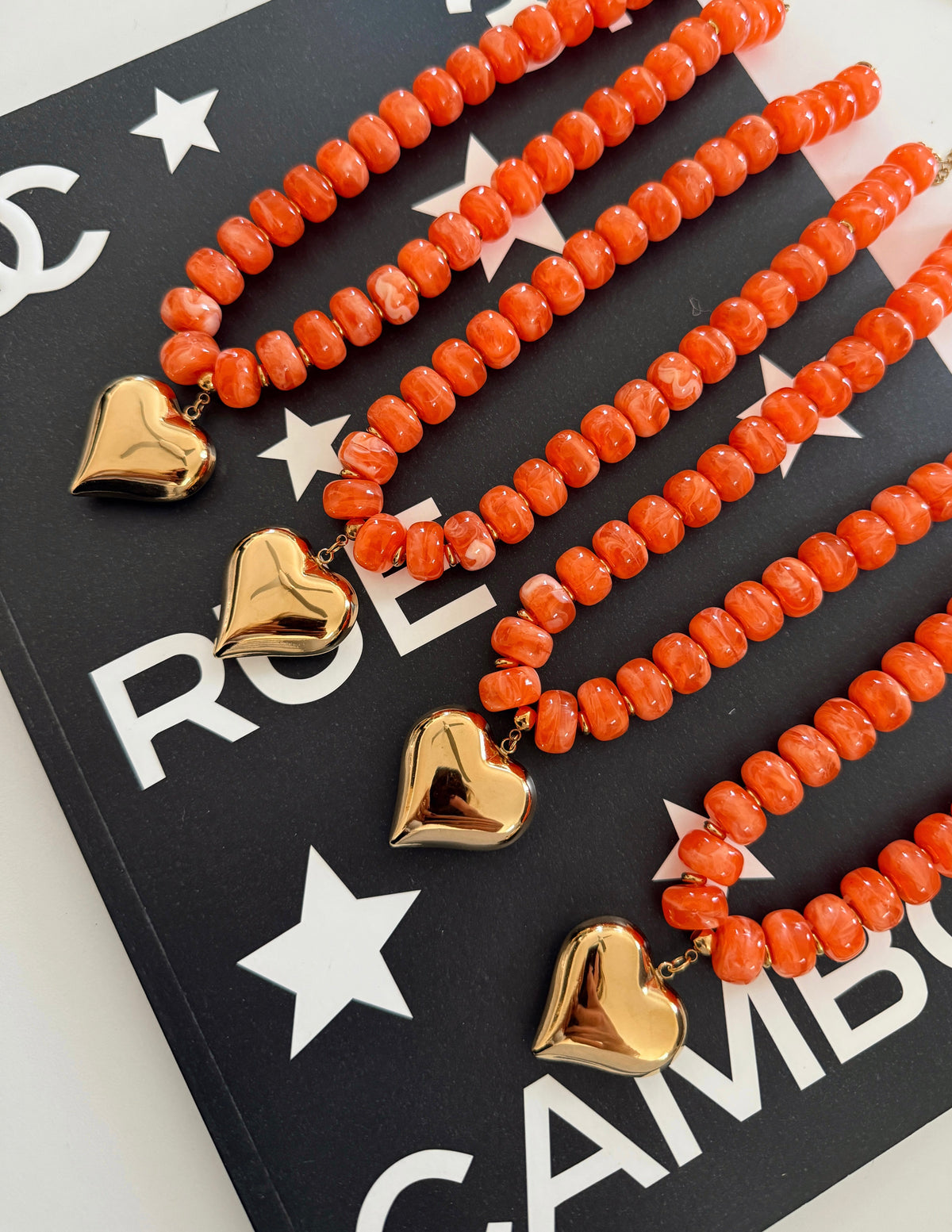 Three coral-orange beaded necklaces with polished gold heart pendants arranged diagonally on a black printed card with white stars and letters.