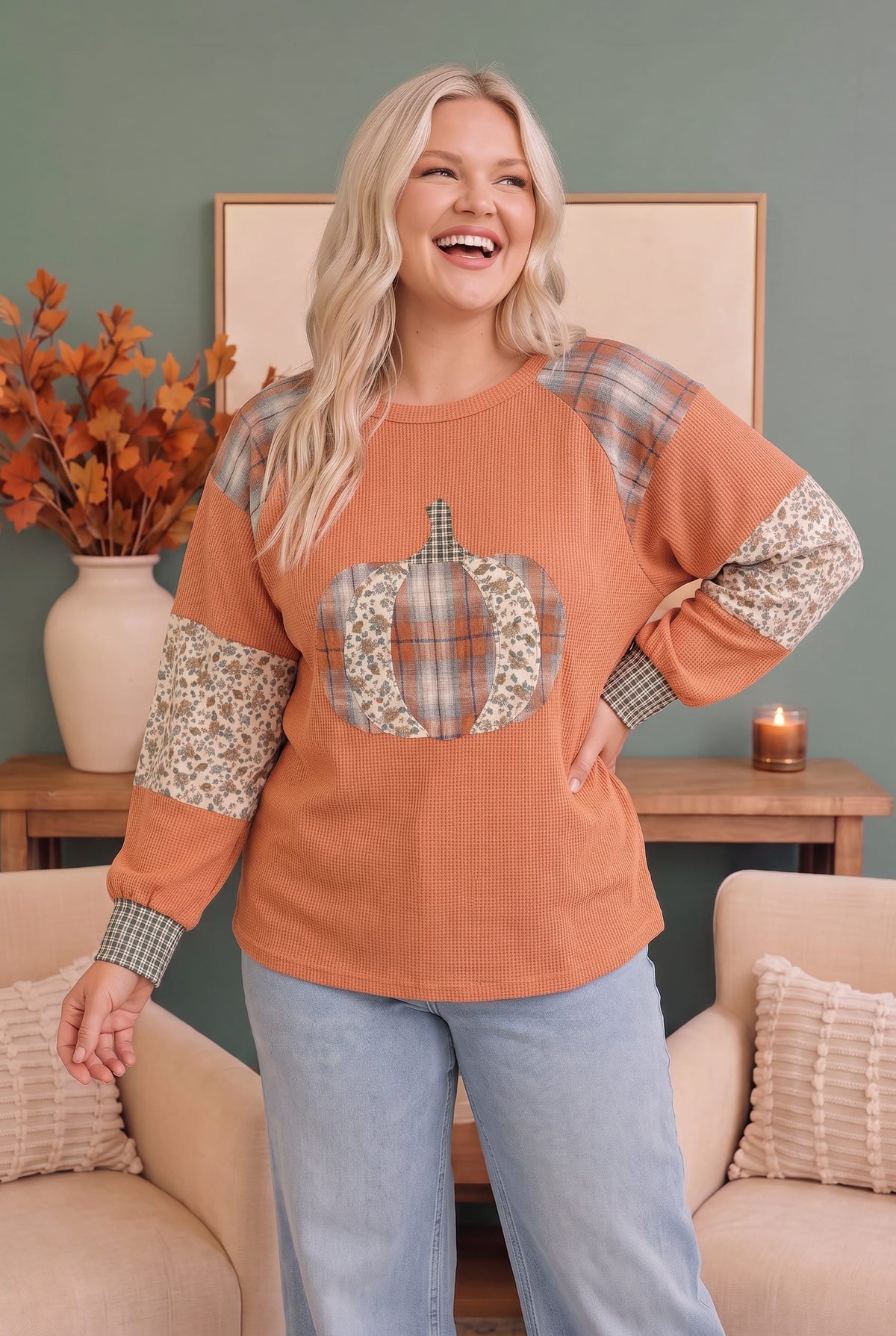 A woman modeling a long-sleeve crewneck sweatshirt in a cozy indoor setting. The sweatshirt is terracotta orange with plaid and floral patchwork details on the shoulders, sleeves, and a pumpkin graphic on the chest.