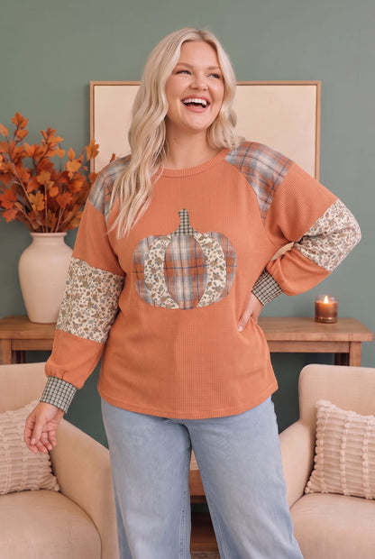A woman modeling a long-sleeve crewneck sweatshirt in a cozy indoor setting. The sweatshirt is terracotta orange with plaid and floral patchwork details on the shoulders, sleeves, and a pumpkin graphic on the chest.