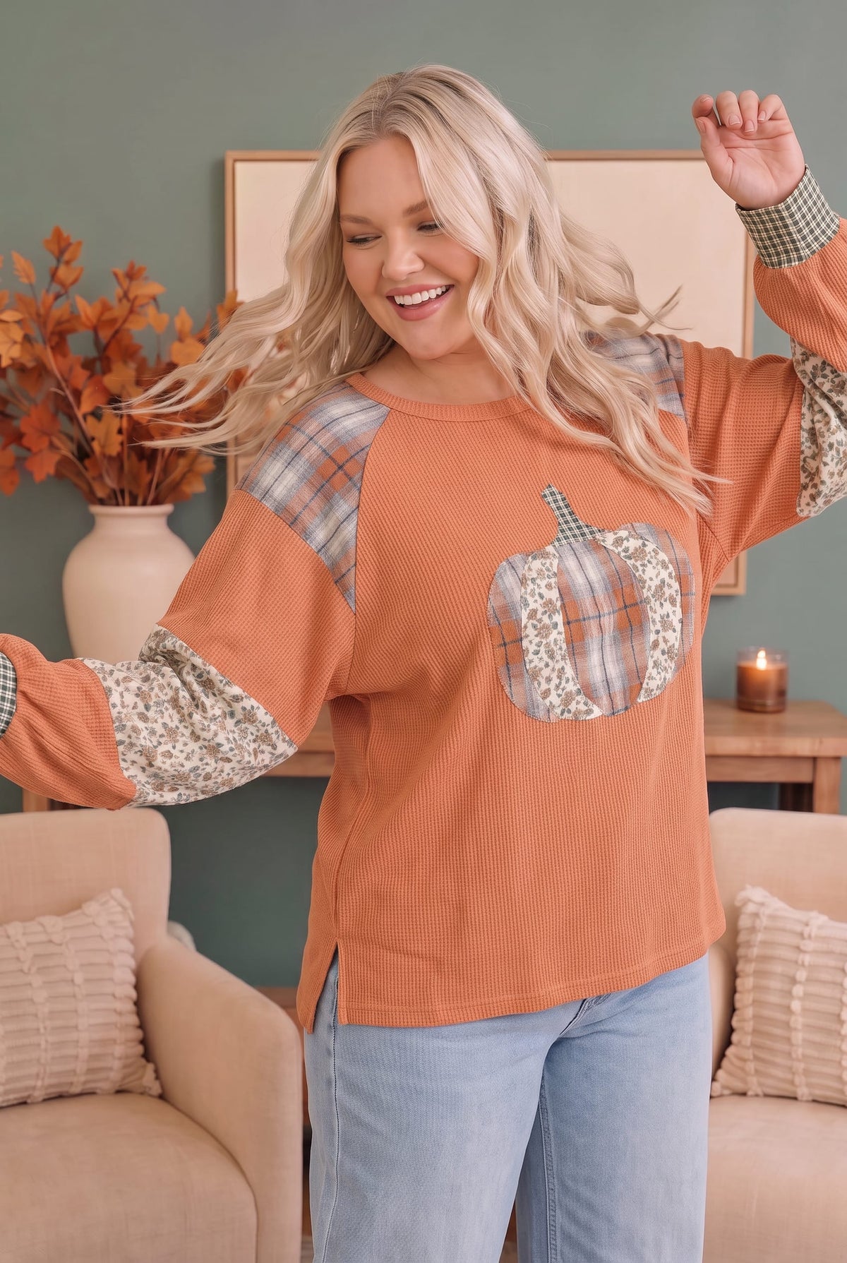 A woman modeling a long-sleeve crewneck top in a warm rust orange color, featuring plaid shoulder and sleeve panels, a floral pocket, and a pumpkin applique on the chest, paired with light blue jeans in a cozy living room setting.