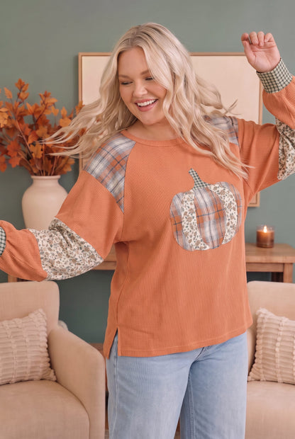 A woman modeling a long-sleeve crewneck top in a warm rust orange color, featuring plaid shoulder and sleeve panels, a floral pocket, and a pumpkin applique on the chest, paired with light blue jeans in a cozy living room setting.
