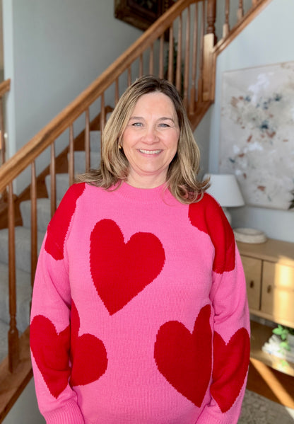 Woman wearing a pink sweater with red heart patterns in a home setting.