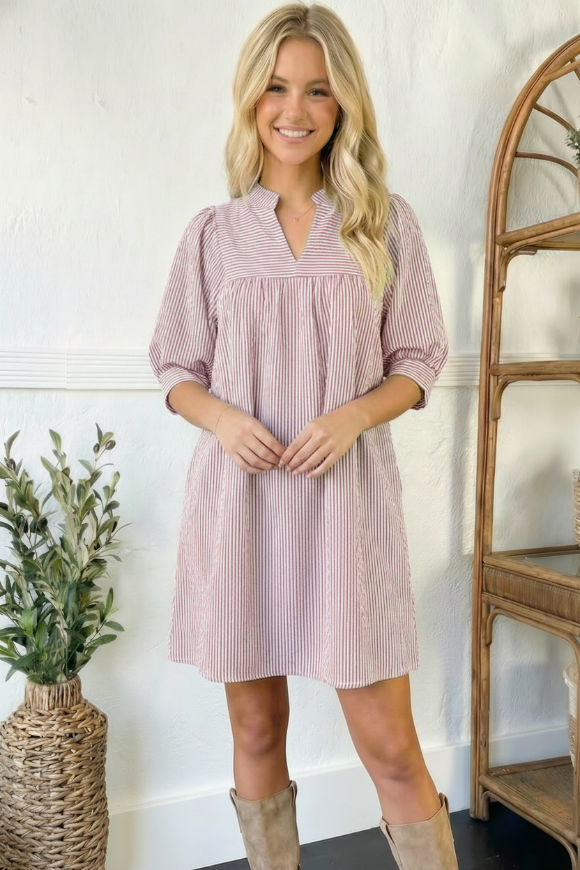 Woman wearing a pink striped dress standing in a room with a plant and wooden shelf.
