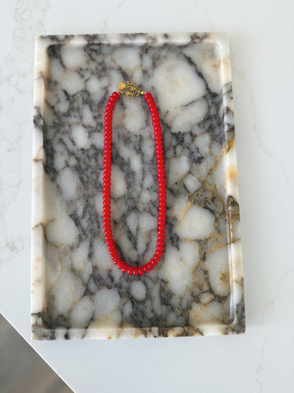 A red beaded necklace with a gold clasp is displayed on a rectangular white and gray marble tray over a white marble surface.