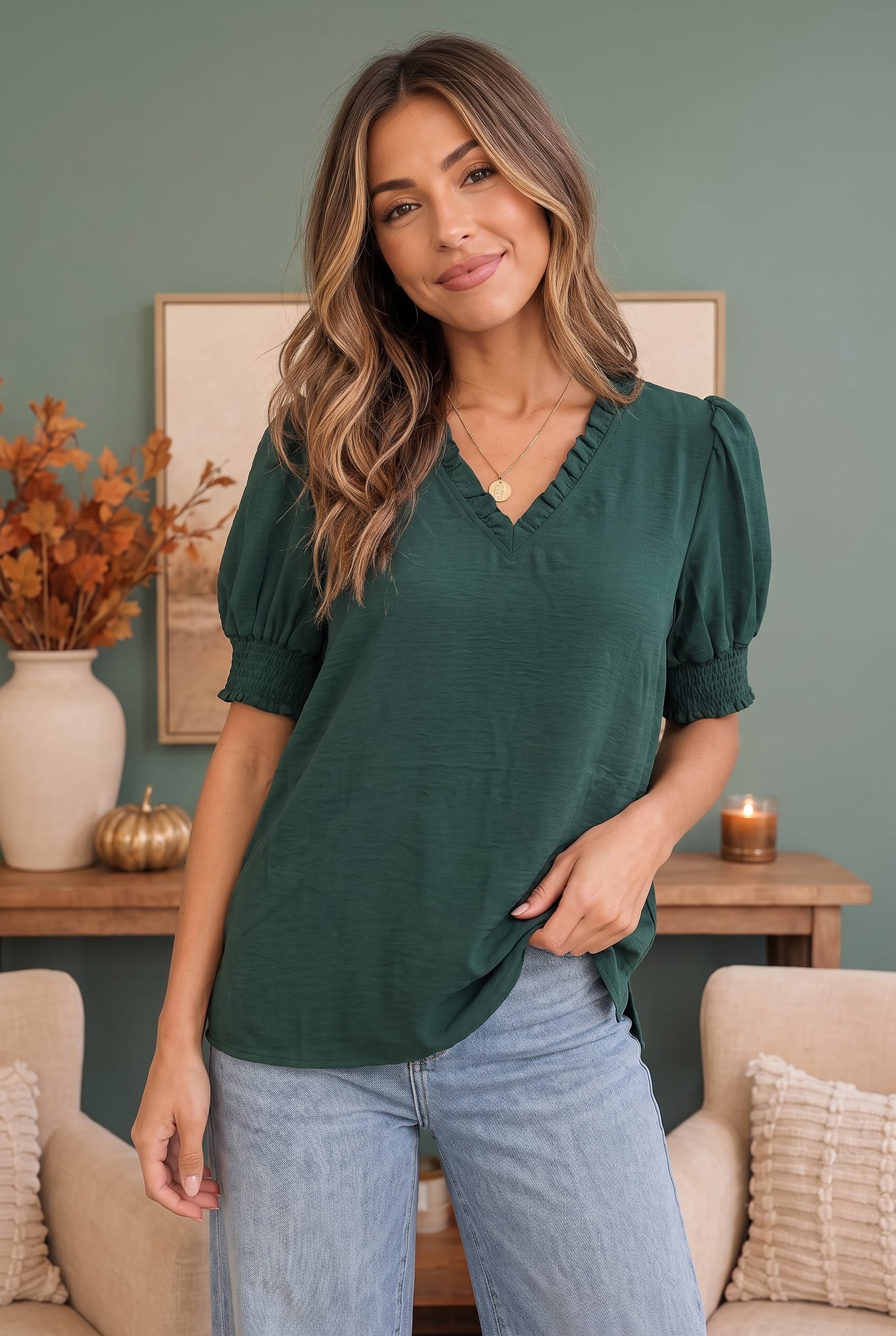 A woman wearing a dark green short-sleeve blouse with puff sleeves and a ruffled V-neckline, paired with light blue jeans, standing in a cozy living room setting.