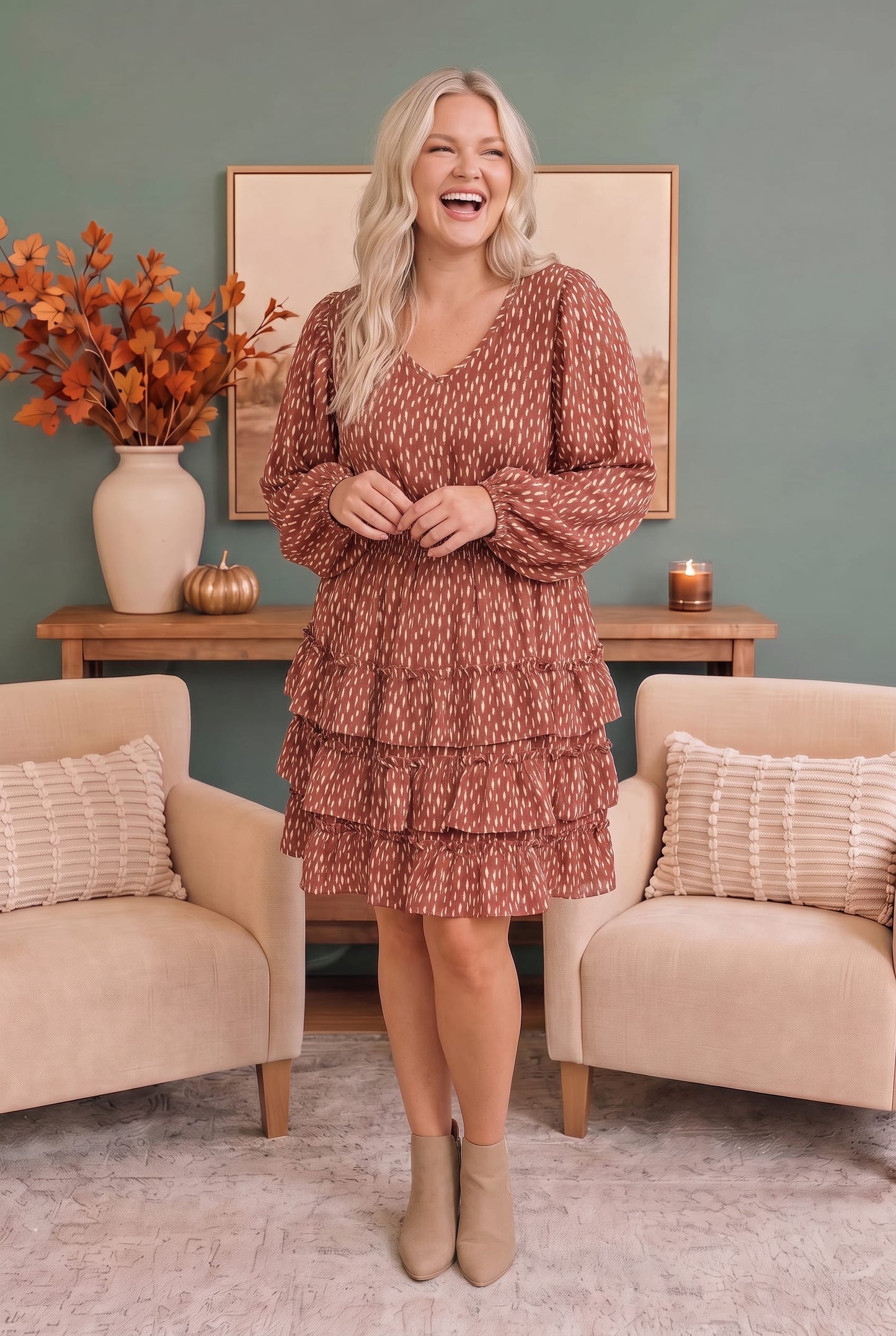 A woman stands in a styled room wearing a rust-colored, tiered mini dress with a white abstract pattern and beige ankle boots.