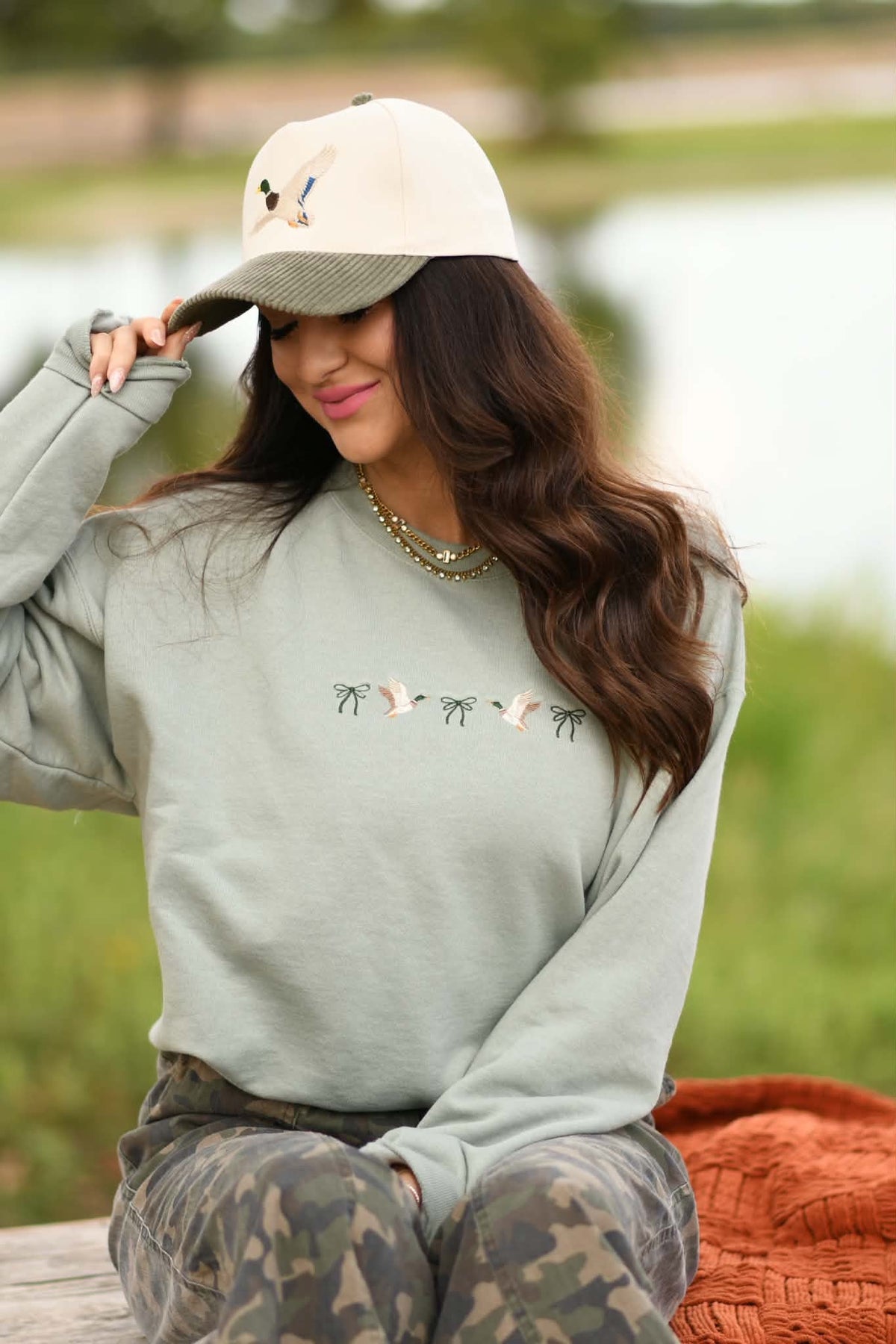A woman wearing a sage green crewneck sweatshirt with embroidered birds, a beige baseball cap, and camouflage pants, sitting outdoors near a lake.