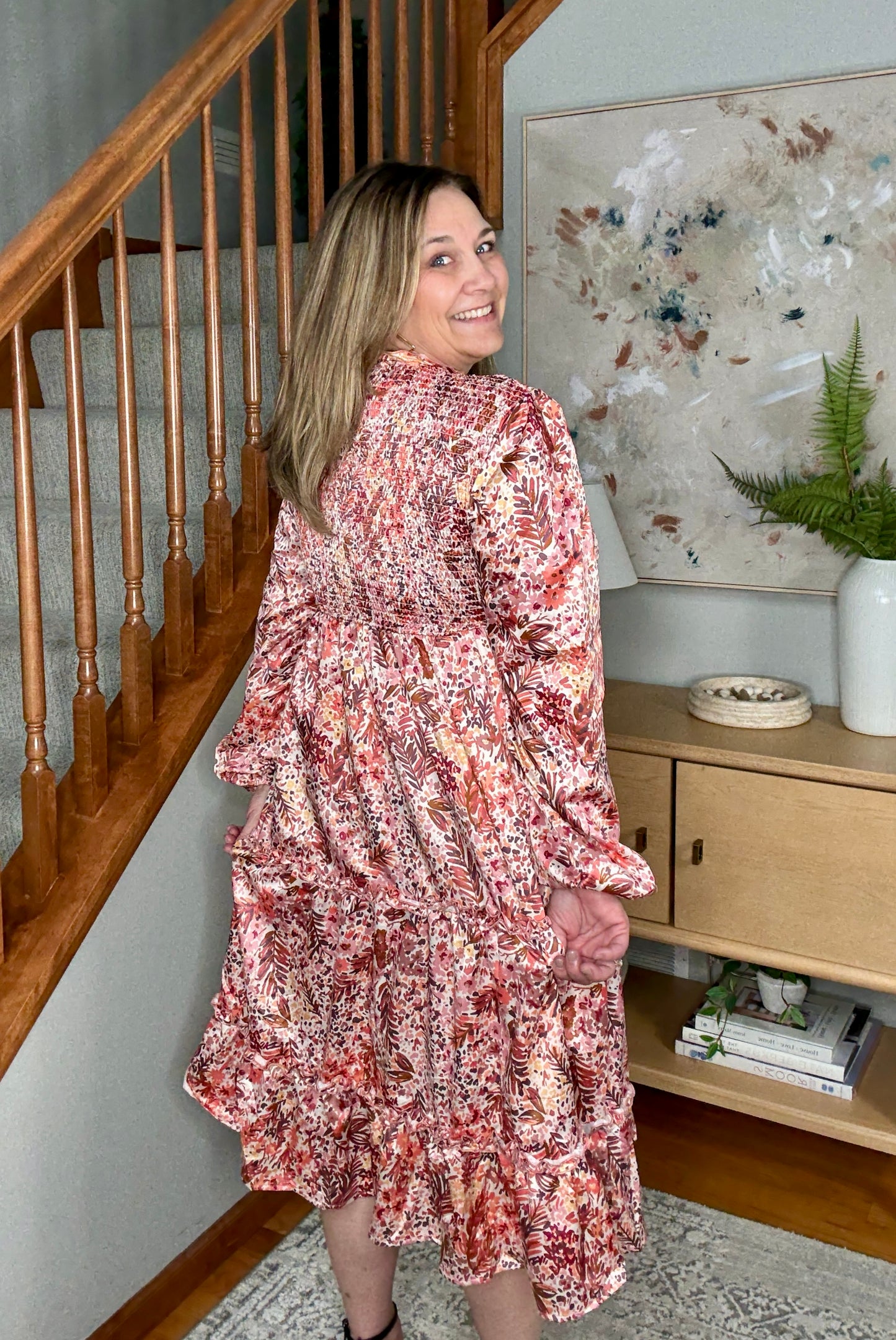Woman in a floral dress standing in a home interior with a staircase and decorative elements.