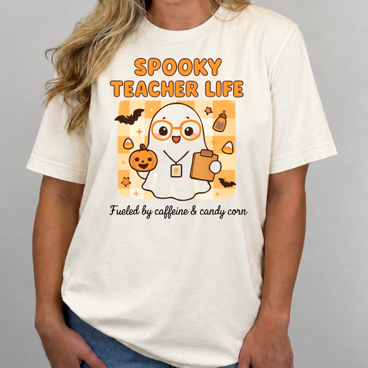 A woman wearing a cream-colored graphic t-shirt with a cartoon ghost teacher design and the text 'SPOOKY TEACHER LIFE' and 'Fueled by caffeine & candy corn'.