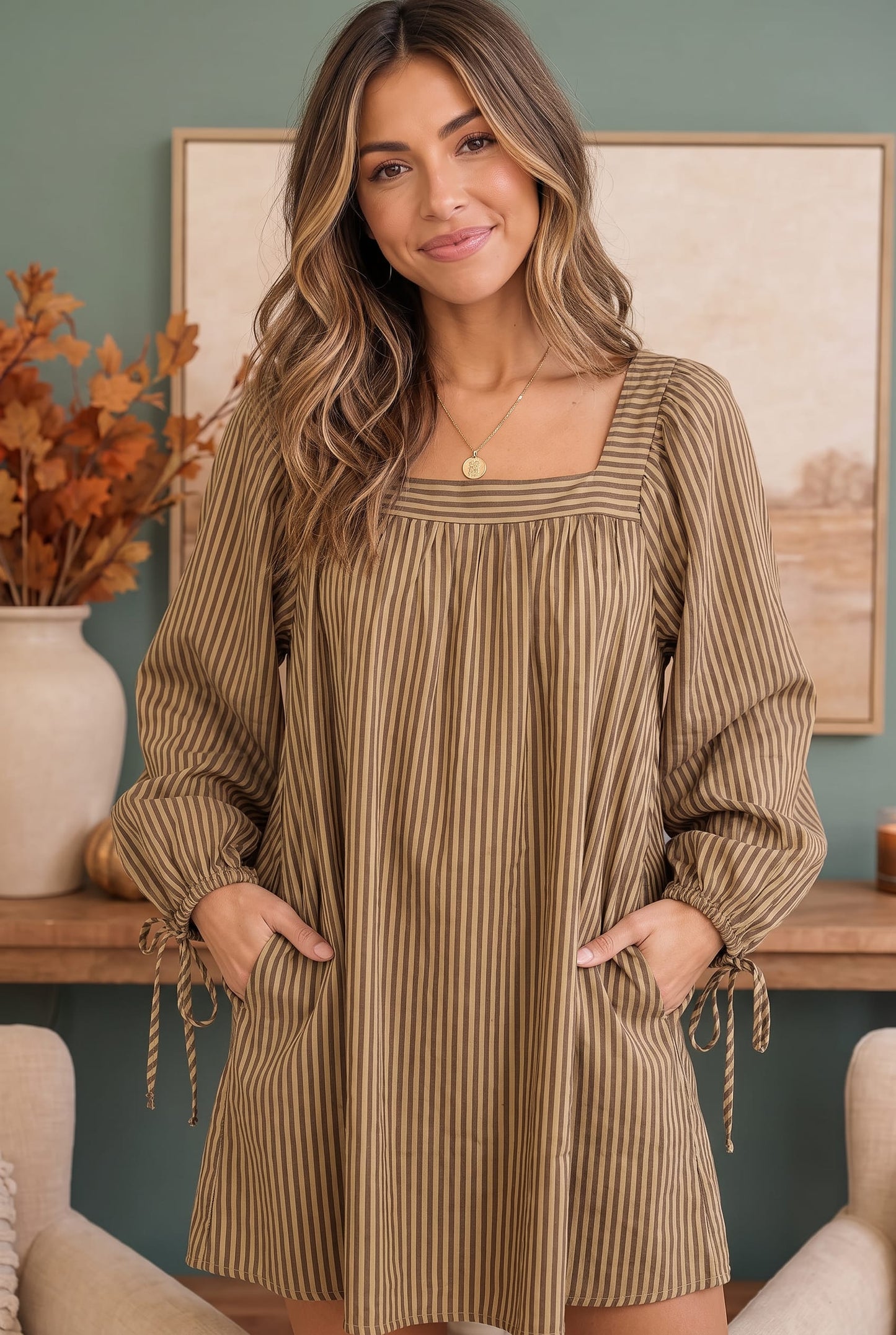 A woman wearing a brown and beige vertically striped long-sleeve tunic dress with a square neckline, front pockets, and tie details on the sleeves, standing in a styled room with a teal wall and framed art.