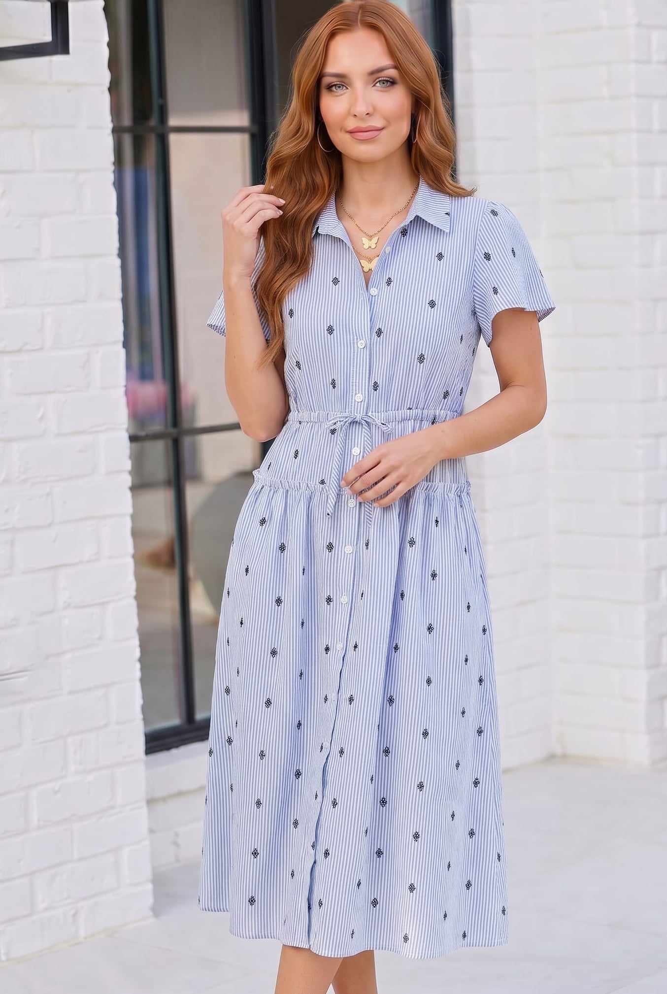 Woman wearing a blue and white striped dress with black polka dots against a white brick wall.