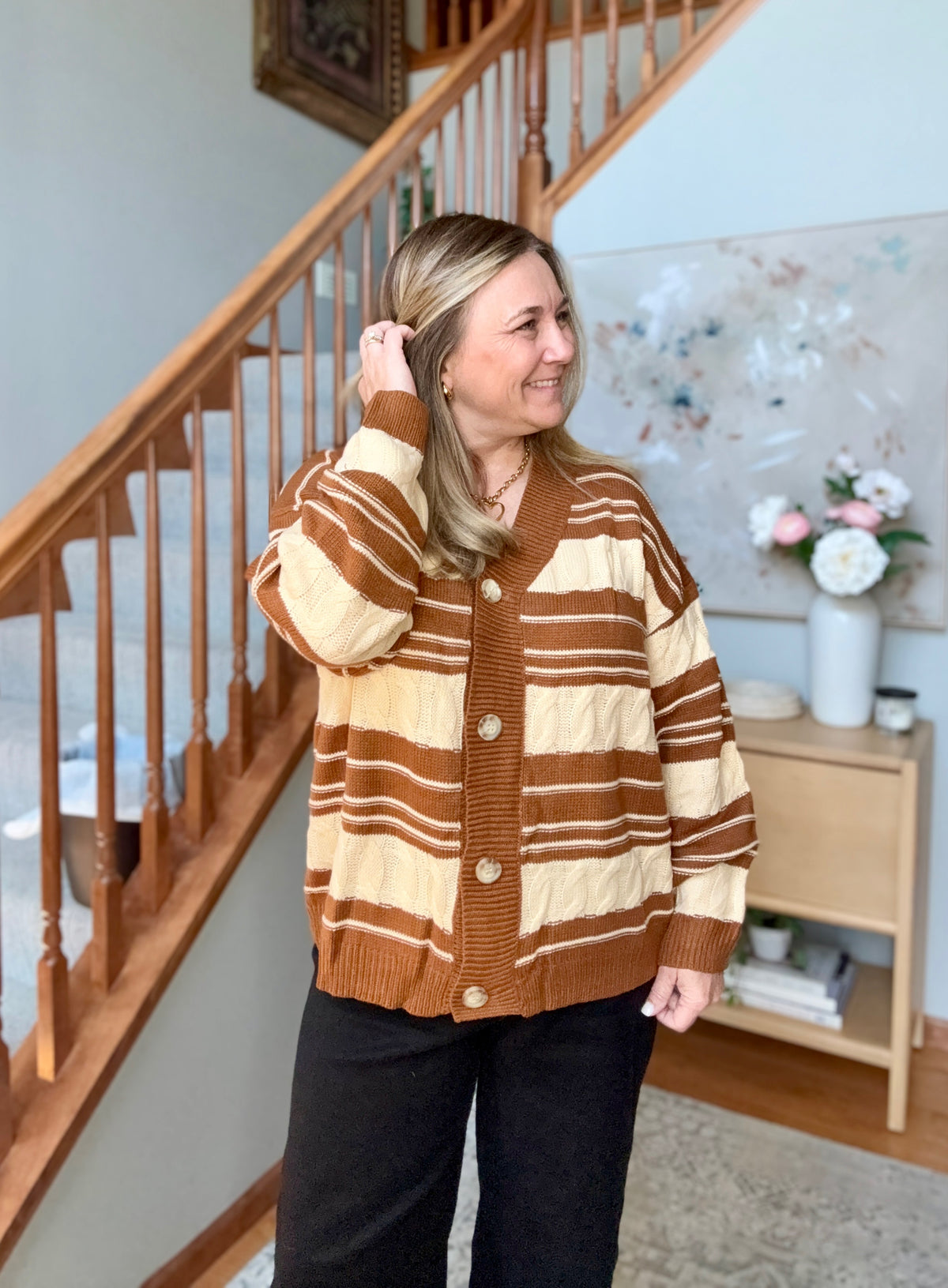 A woman wearing a brown and cream striped knit cardigan sweater with buttons, standing in a home interior next to a wooden staircase.