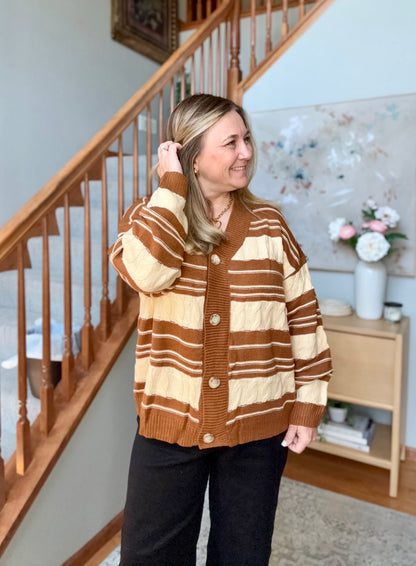 A woman wearing a brown and cream striped knit cardigan sweater with buttons, standing in a home interior next to a wooden staircase.