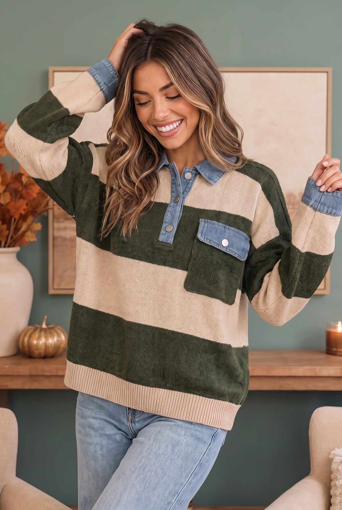 A woman wearing a striped knit sweater with dark green, beige, and light blue denim accents, paired with light blue jeans, in a cozy indoor setting.