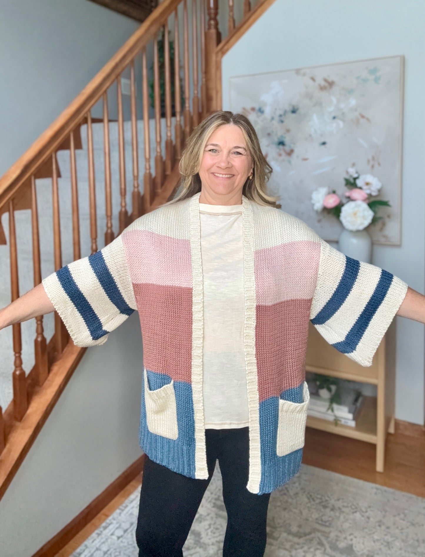 A woman wearing a chunky knit color-block cardigan with cream, dusty pink, navy blue, and light blue panels, standing in a home interior with a staircase in the background.