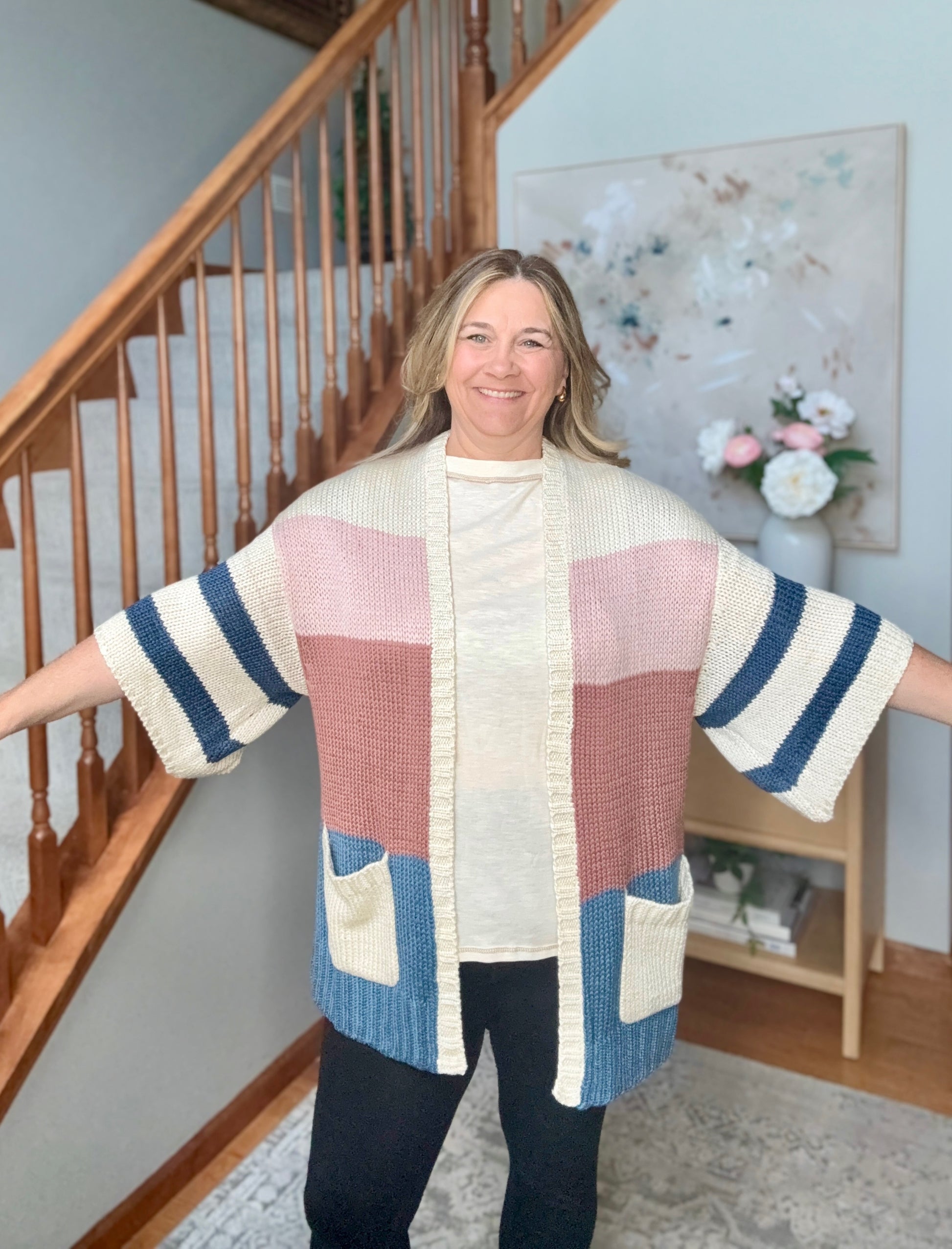 A woman wearing a chunky knit color-block cardigan with cream, dusty pink, navy blue, and light blue panels, standing in a home interior with a staircase in the background.