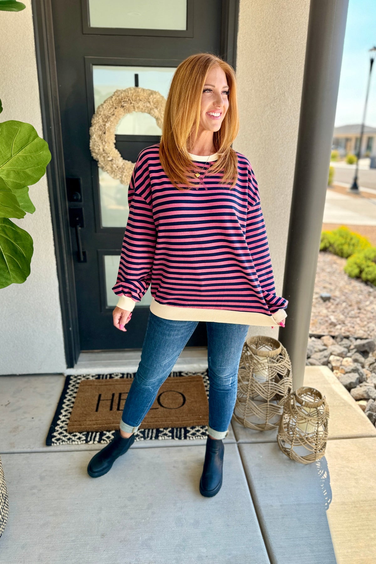 Woman standing on a porch wearing a striped sweater and blue jeans, with a front door and doormat in the background.