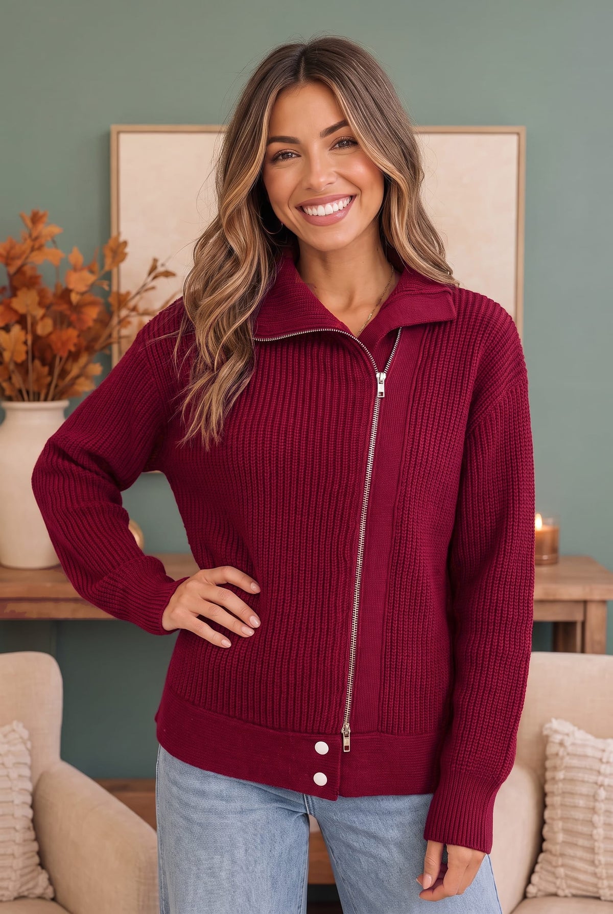 A smiling woman wearing a ribbed knit zip-front sweater in deep burgundy with a stand collar and light blue jeans, standing indoors in a living room setting.