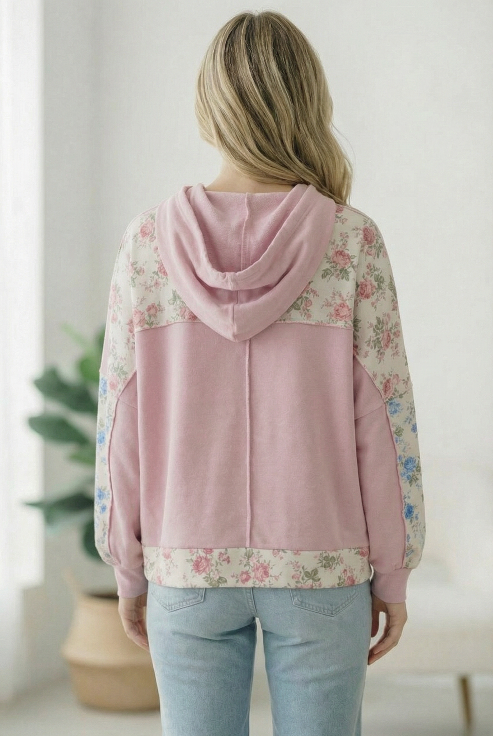 Person wearing a pink hoodie with floral sleeves in a room with a plant.