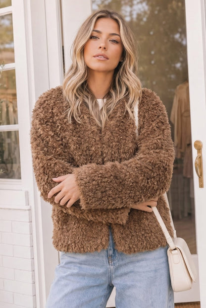 A woman wearing a brown curly faux fur cardigan, light blue jeans, and carrying a cream shoulder bag, standing in front of a white door.