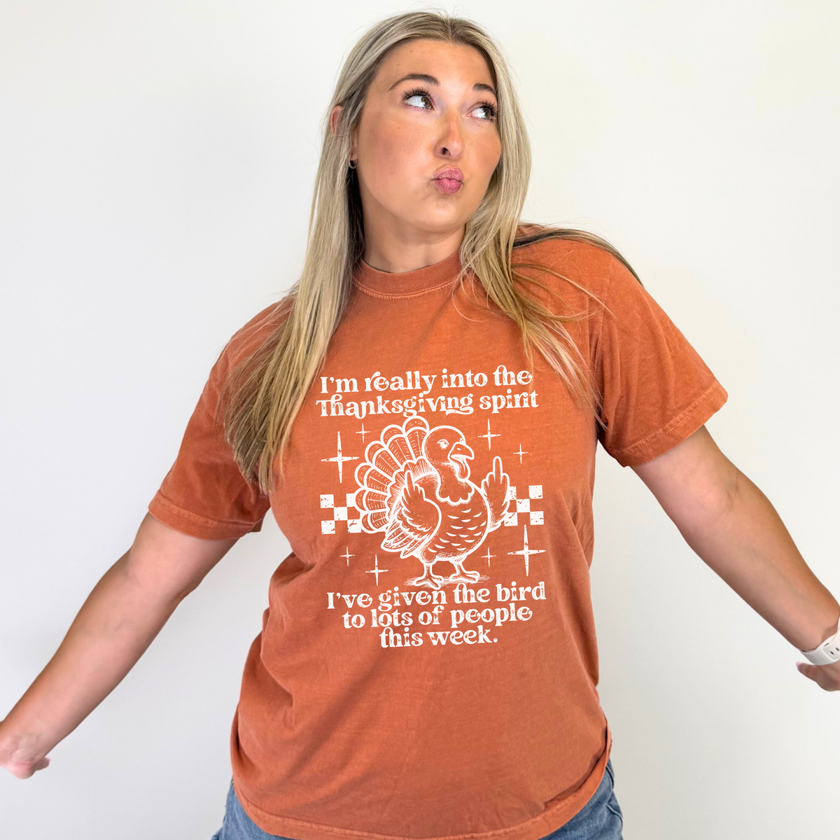 A woman wearing a burnt orange graphic t-shirt with a white turkey illustration and text that reads, 'I'm really into the Thanksgiving spirit I've given the bird to lots of people this week.' She is posing against a plain white wall.