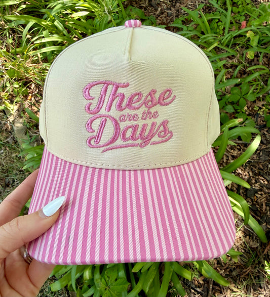A hand holds a baseball cap outdoors with green foliage in the background. The cap has a cream crown with pink embroidered text reading 'These are the Days' and a pink-and-white vertically striped brim.