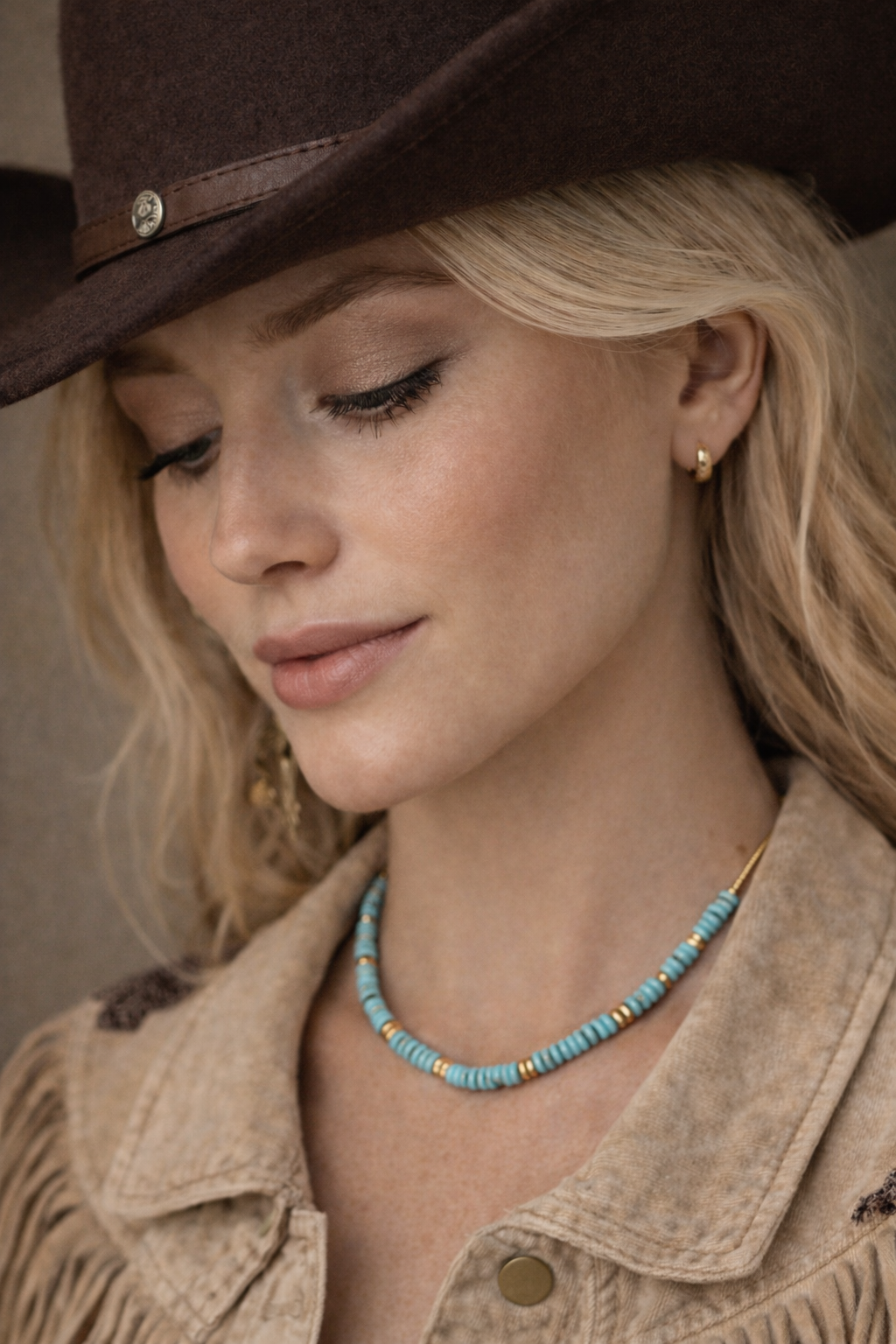 Woman wearing a brown cowboy hat and beige jacket with a blue beaded necklace.