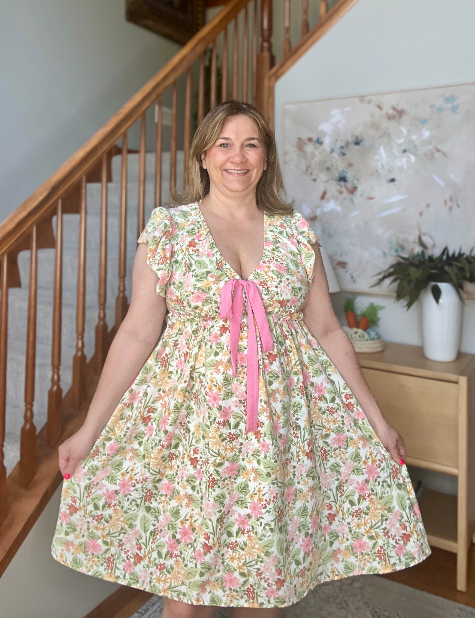 Woman wearing a floral dress with a pink ribbon standing in a home setting.