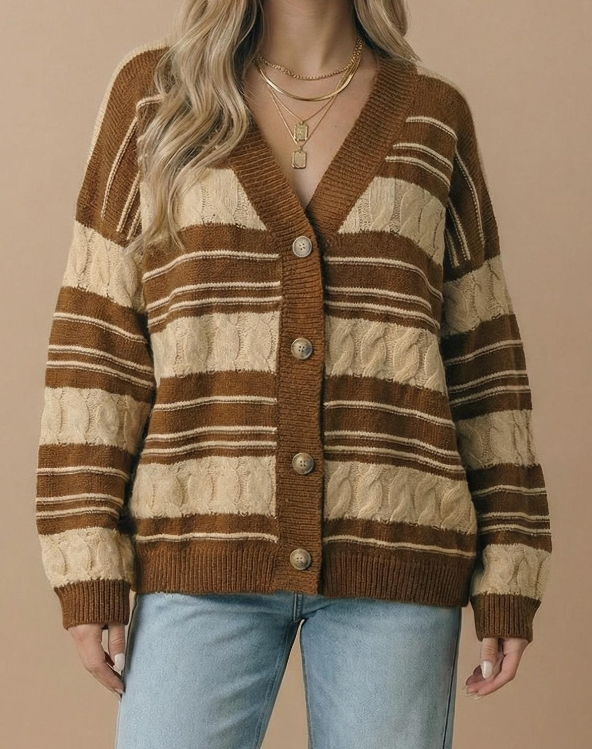 A woman wearing a brown and beige striped cable-knit cardigan sweater with button-front closure, paired with light blue jeans, against a solid beige background.