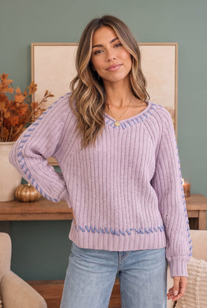 A woman wearing a lavender ribbed knit sweater with blue patterned sleeve and hem details, paired with light blue jeans, standing in a styled indoor setting.