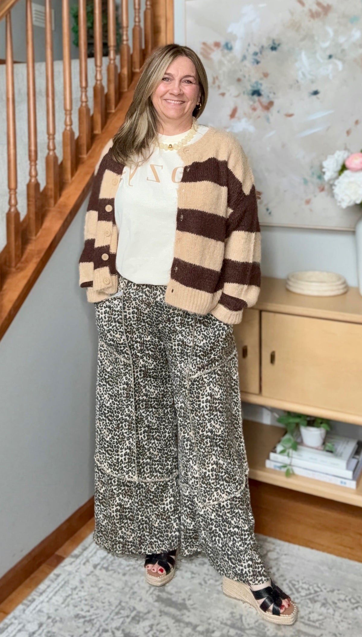 A woman stands in a home interior wearing a cream and brown striped knit cardigan, a cream top, wide-leg patterned pants, and black slide sandals. A staircase, cabinet, and framed wall art are visible in the background.