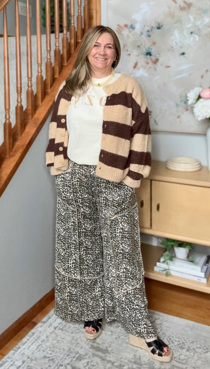 A woman stands in a home interior wearing a cream and brown striped knit cardigan, a cream top, wide-leg patterned pants, and black slide sandals. A staircase, cabinet, and framed wall art are visible in the background.