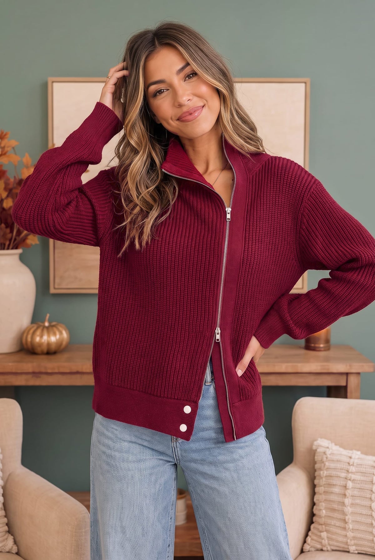 A woman wearing a ribbed knit zip-up jacket in deep burgundy with light blue jeans, standing in a cozy living room setting with a green wall and framed art in the background.