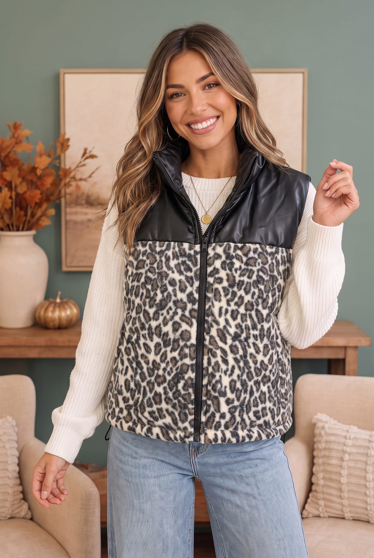 A woman modeling a sleeveless zip-front vest with a black faux-leather yoke and a beige, black, and gray leopard print lower body, worn over a cream ribbed sweater and light blue jeans in a styled indoor setting.