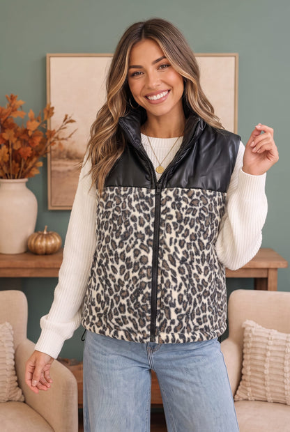 A woman modeling a sleeveless zip-front vest with a black faux-leather yoke and a beige, black, and gray leopard print lower body, worn over a cream ribbed sweater and light blue jeans in a styled indoor setting.